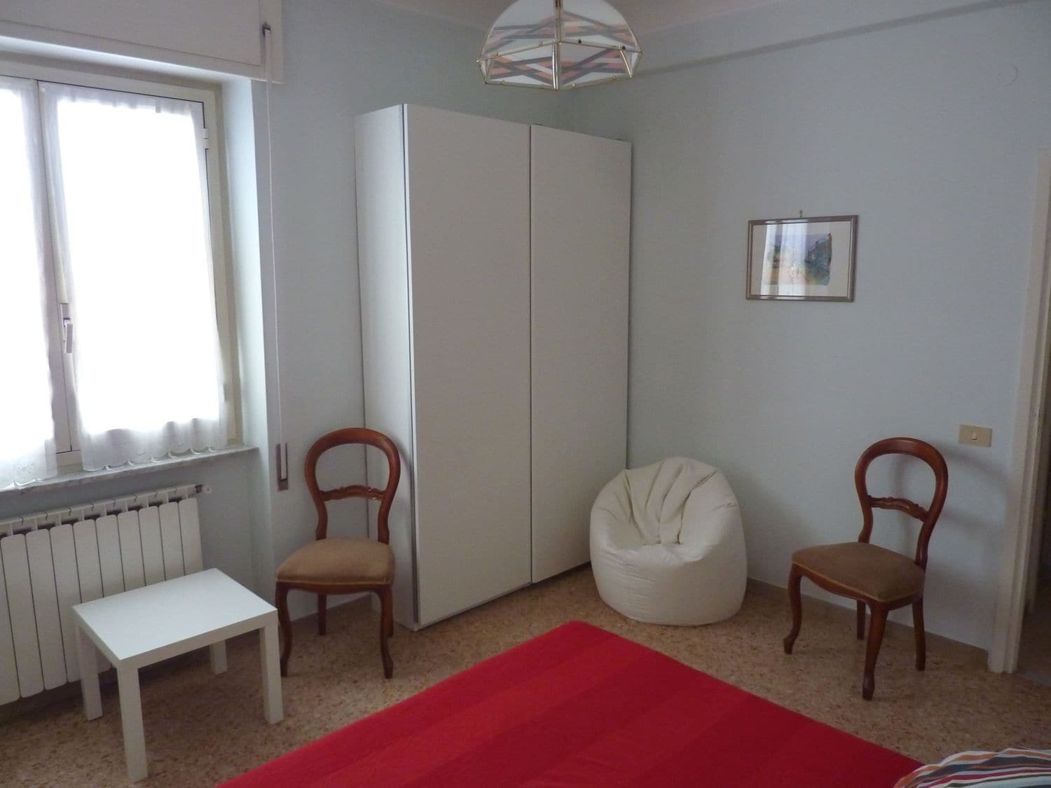 Une pièce avec un radiateur blanc, une armoire blanche, un fauteuil en forme de poire blanc, deux chaises en bois, un tapis rouge et un tableau encadré au mur.