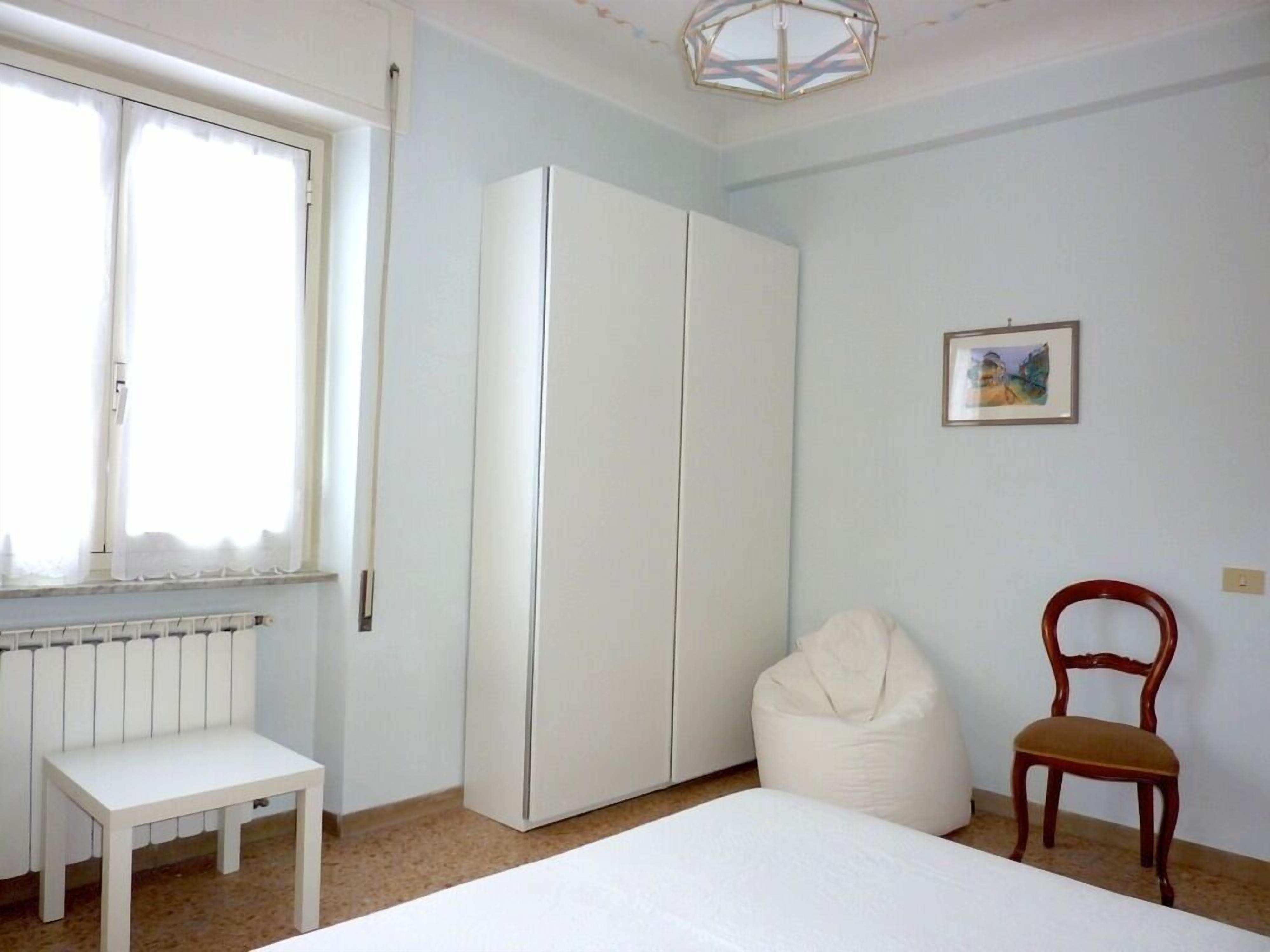 Un dormitorio con cama, armario blanco, radiador, ventana con cortinas, una silla y una pequeña mesa.