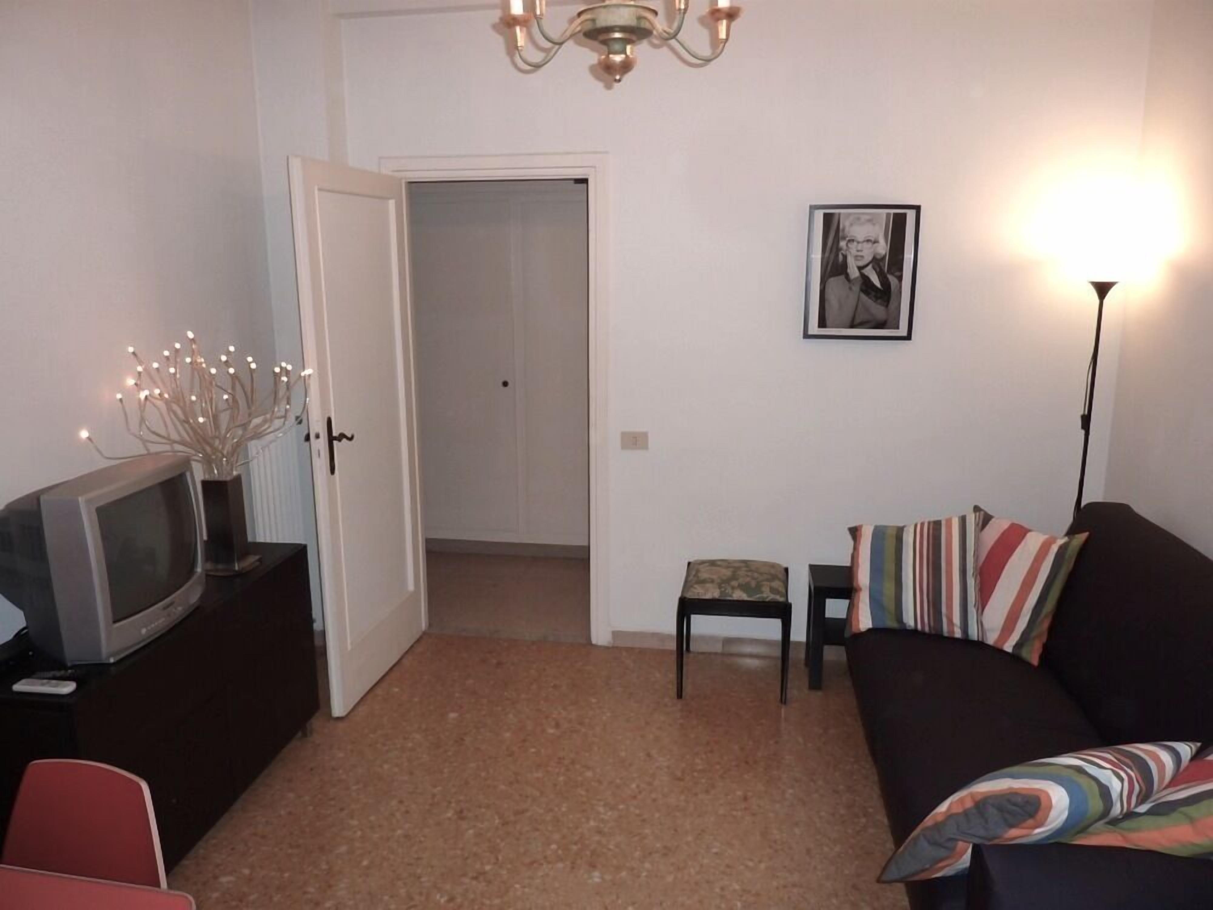A living room with a television on a cabinet, a sofa with striped pillows, a small table with a chair, and a door leading to another room.