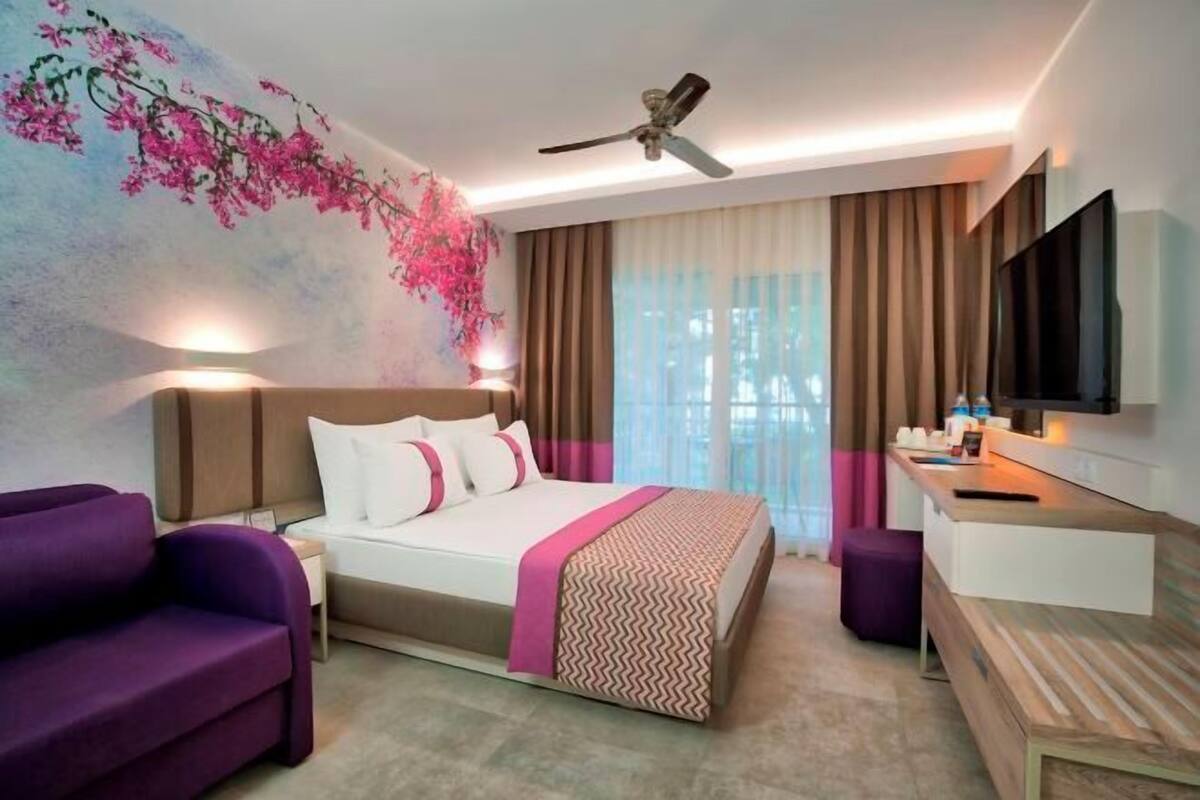A hotel room with a bed, a desk, a television, a ceiling fan, and a window with curtains.