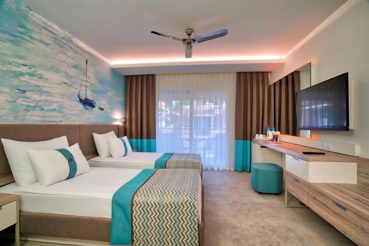 A hotel room with two beds, a TV, a ceiling fan, and a large window with curtains.