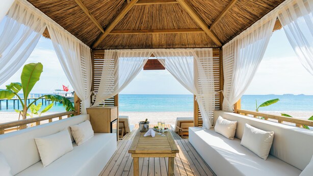 A beachside cabana with a wooden table, white cushioned seating, and a view of the ocean. - Pirates Beach Club - All Inclusive (Kemer)