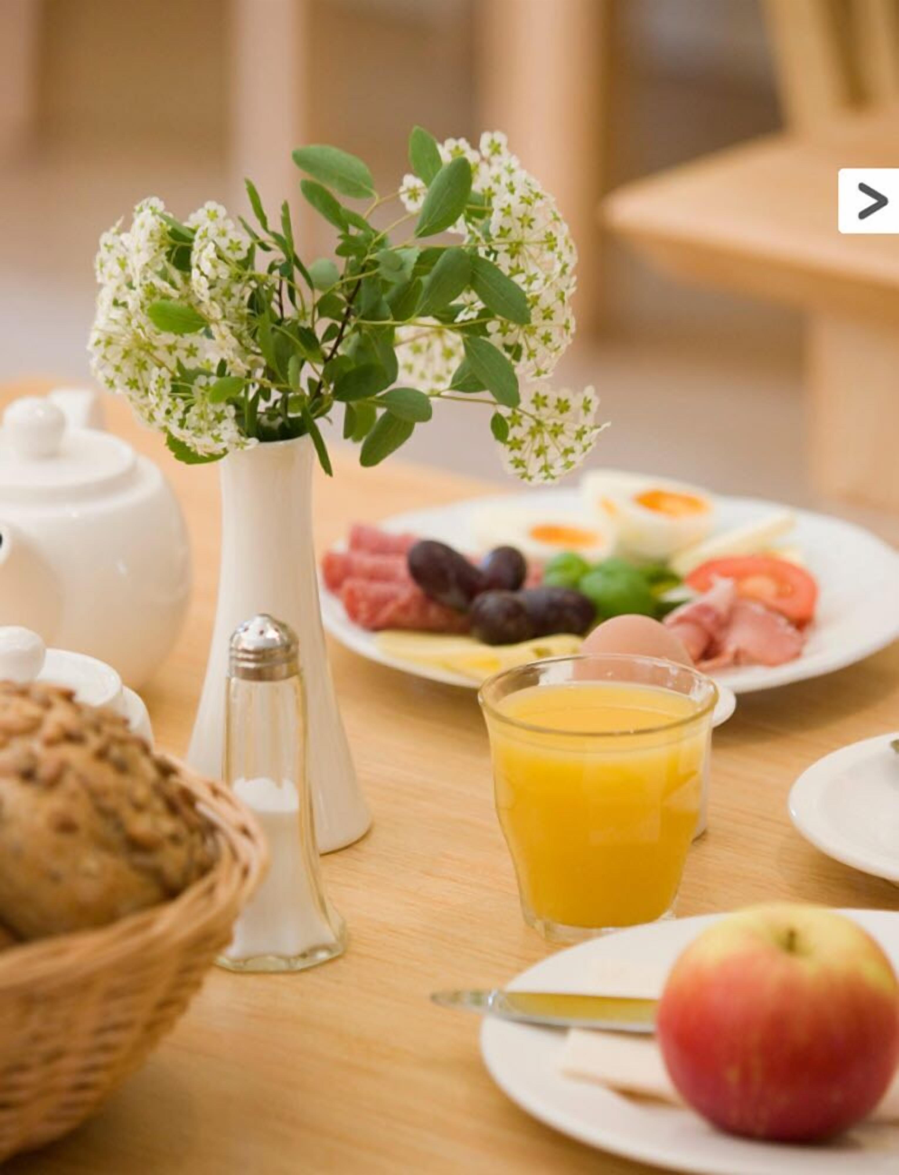 Een ontbijttafel met een witte theepot, een zoutvaatje, een glas jus d'orange, een bord met eieren, fruit en een plak ham.