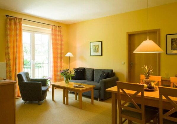 Una sala de estar con sofá, sillón, mesa de centro y mesa de comedor. Hay una ventana con cortinas, una lámpara y un cuadro en la pared.