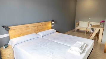 A neatly made bed with white linens and a wooden headboard, a small wooden stool with a bottle, a table with a chair, and a wall-mounted lamp.
