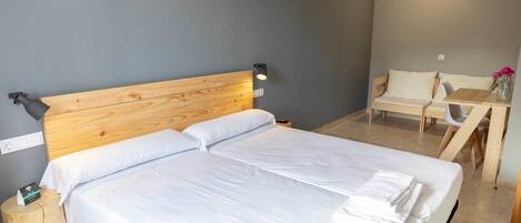 Una cama bien hecha con sábanas blancas y un cabecero de madera, un taburete pequeño de madera con una botella, una mesa con una silla y una lámpara fijada a la pared.