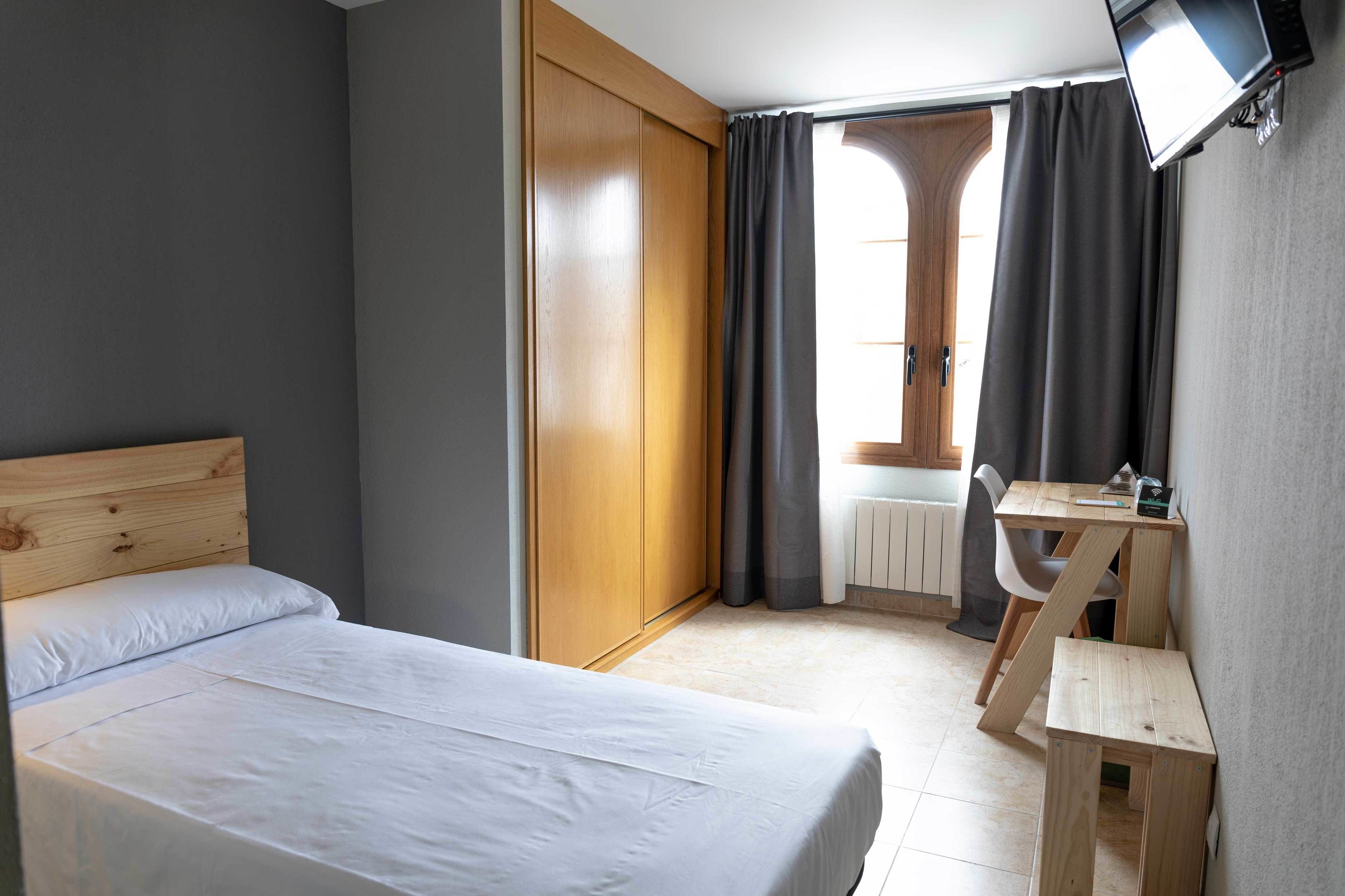 A bedroom with a bed, a wooden wardrobe, a desk with a chair, and a television mounted on the wall.