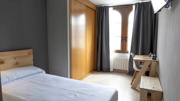 A bedroom with a bed, a wooden wardrobe, a desk with a chair, and a television mounted on the wall.