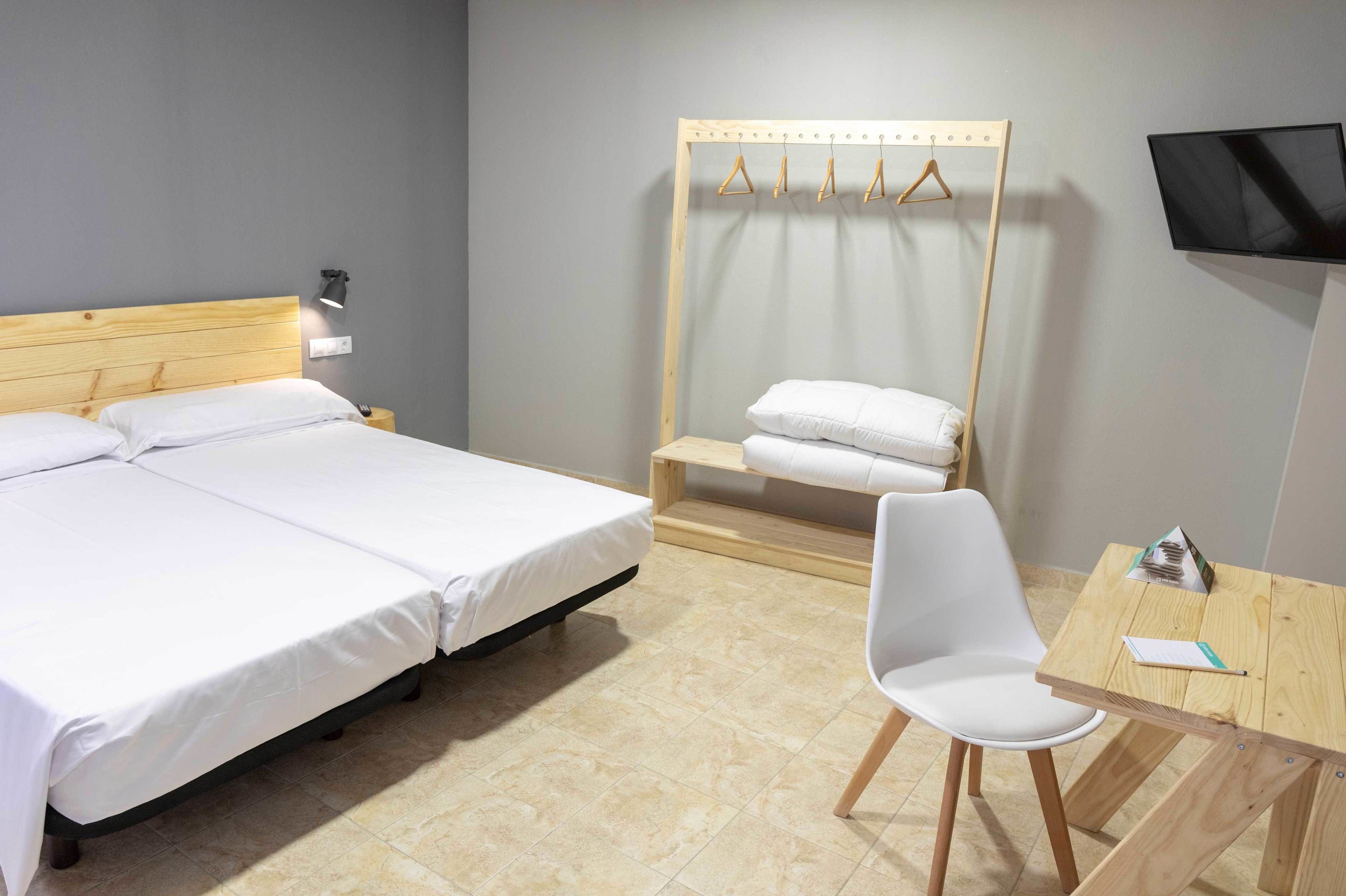 A hotel room with a double bed, a wooden bedside table, a chair, a TV, and a wooden shelf with hanging clothes.