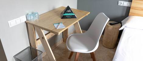 A compact hotel room with a wooden desk, a white chair, a small side table, a bed with white linens, and a wall-mounted lamp.