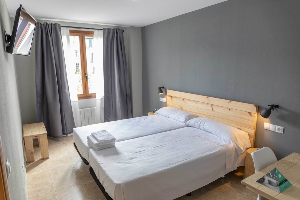Une chambre d’hôtel avec un grand lit, une tête de lit en bois, une table de chevet, une petite commode, un petit tabouret en bois, une fenêtre avec des rideaux et une lampe fixée au mur.