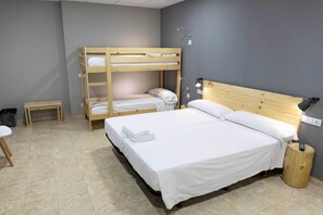 Quarto com cama beliche, uma cama de solteiro, uma mesinha, uma cadeira, abajur e lixeira.