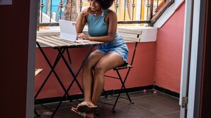 Una persona sentada a una mesa con una computadora portátil en un balcón, con una palmera y edificios al fondo.