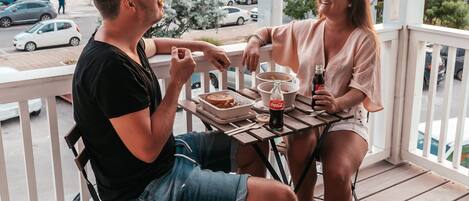Un hombre y una mujer sentados a una mesa en una terraza de madera con barandilla, con vista a una calle con edificios de colores.
