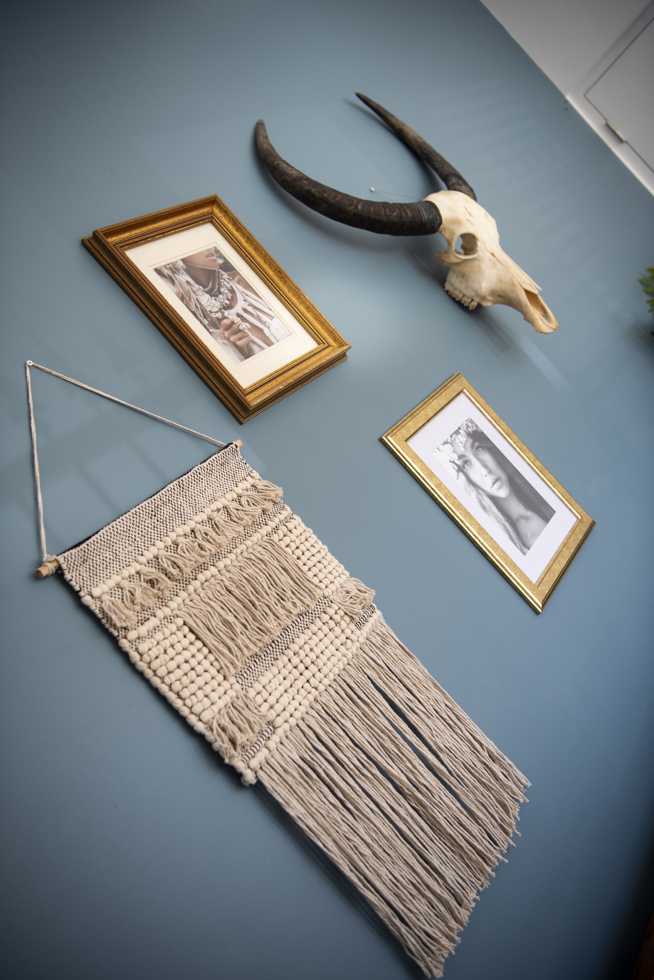 Un tapiz de macramé, un cráneo y dos fotografías enmarcadas sobre una pared azul.