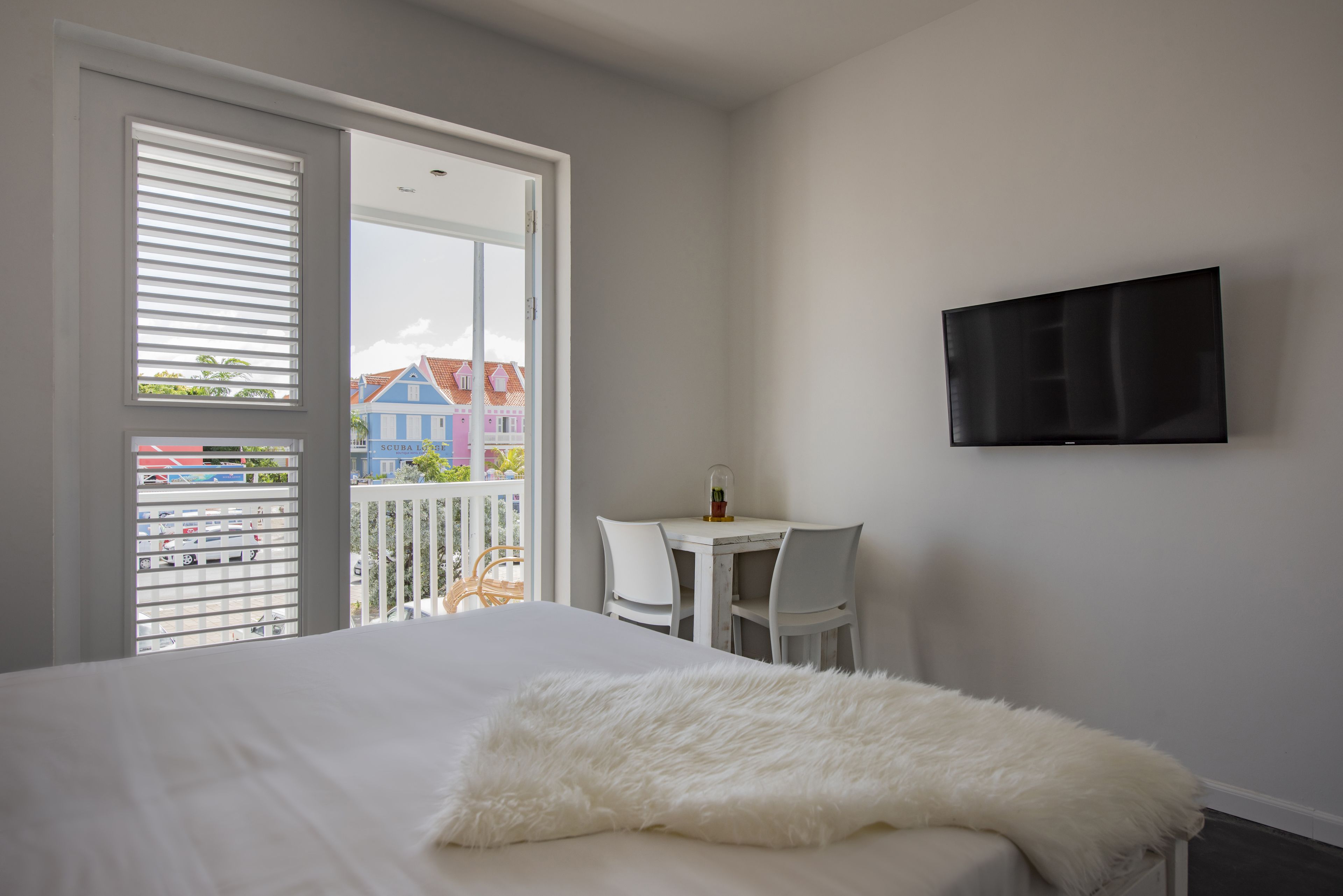 Une chambre avec un lit, une couverture blanche et moelleuse, un téléviseur fixé au mur, une petite table avec des chaises et un balcon donnant sur des maisons.