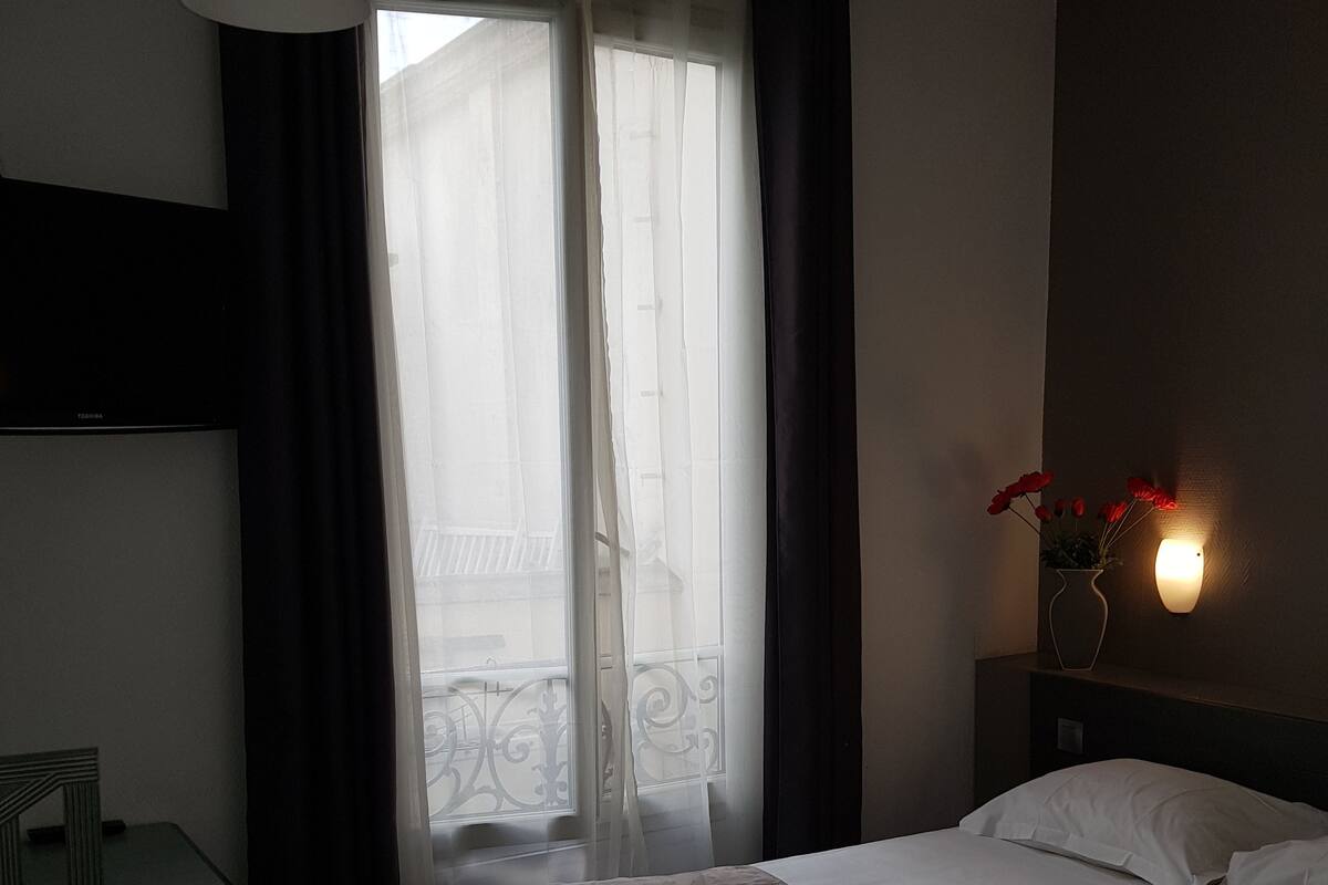 A hotel room with a bed, a desk, a chair, a window with curtains, and a wall-mounted light fixture.