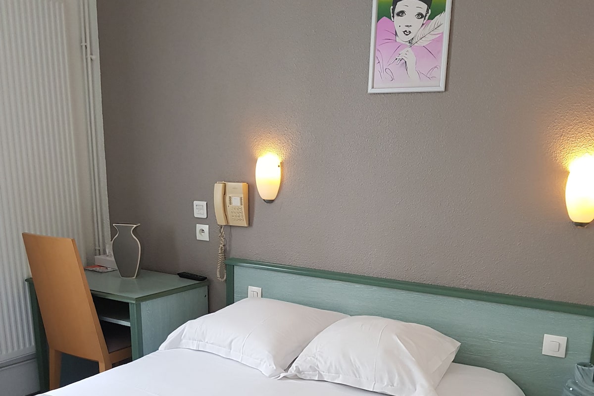 A hotel room with a bed, two bedside tables, a chair, a vase, and a painting on the wall.