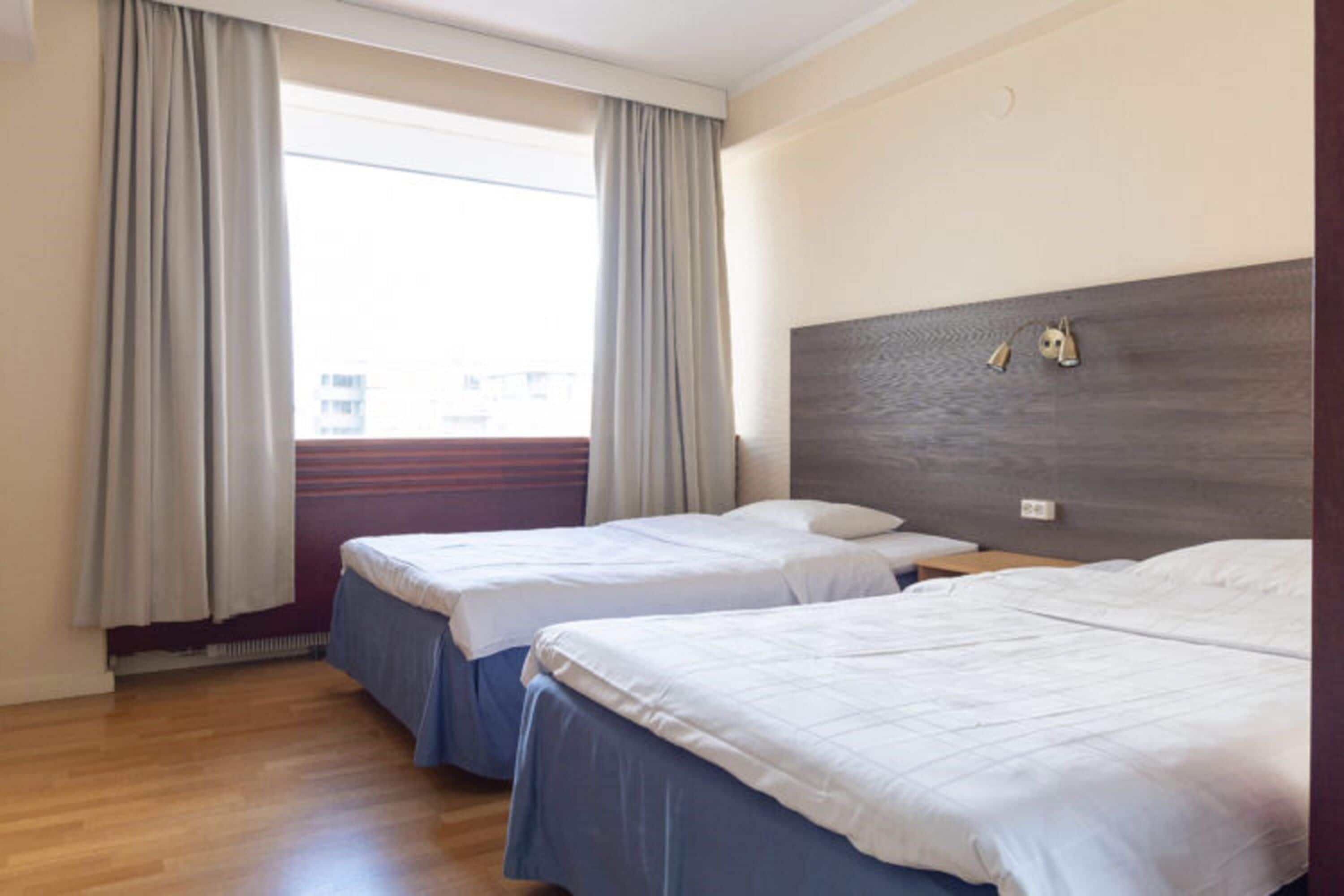 Ein Hotelzimmer mit zwei Betten, einem großen Fenster mit Vorhängen, einem Holzboden und einem Kopfteil mit Wandlampen.