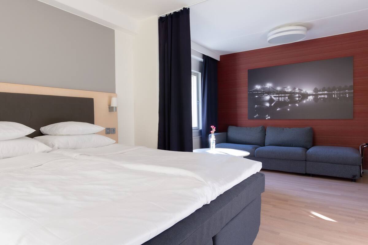 A hotel room with a bed, a gray sofa, a nightstand, and a wall-mounted picture.
