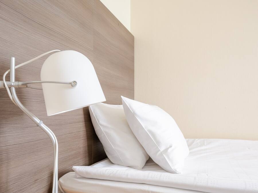 A neatly made bed with white linens and pillows, a modern wall with a wooden panel, and a white bedside lamp.