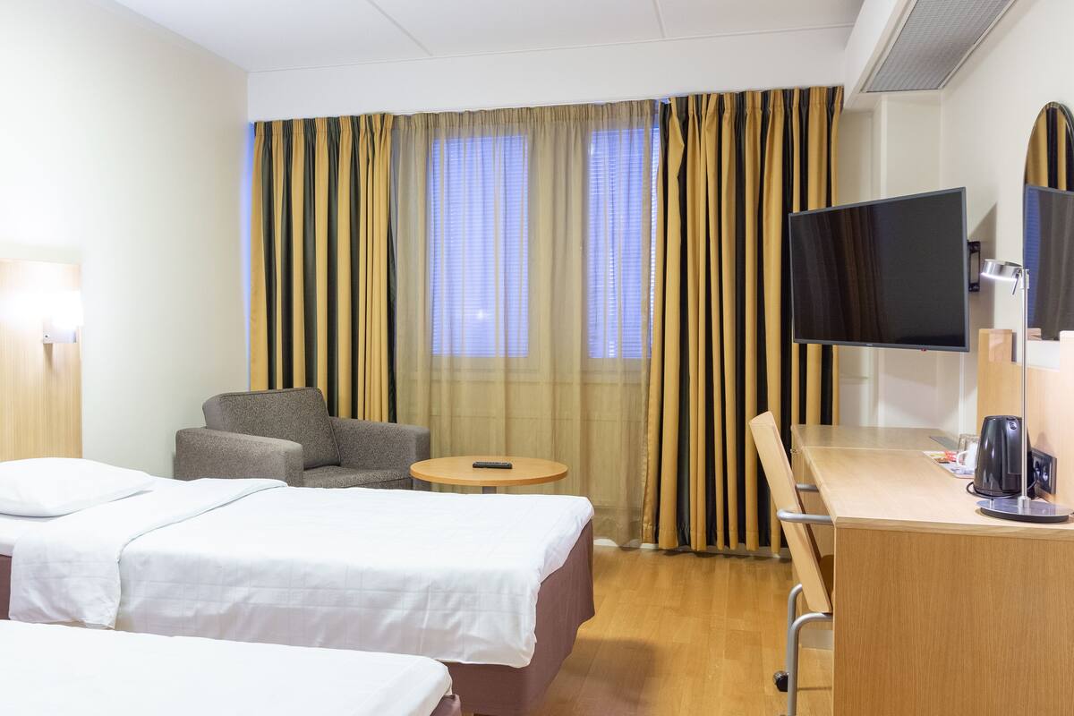 A hotel room with two beds, a desk, a chair, a TV, and a window with curtains.