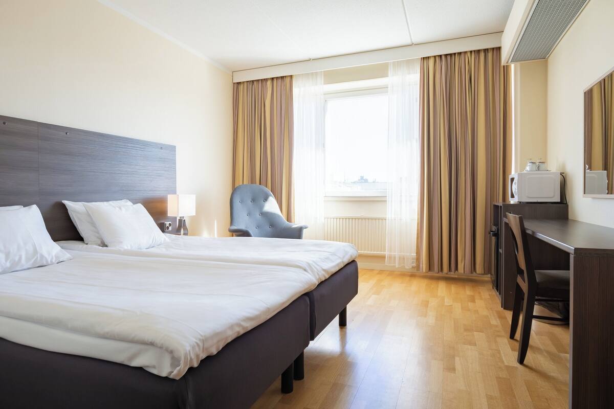 A hotel room with a large bed, a desk, a chair, and a window with curtains.
