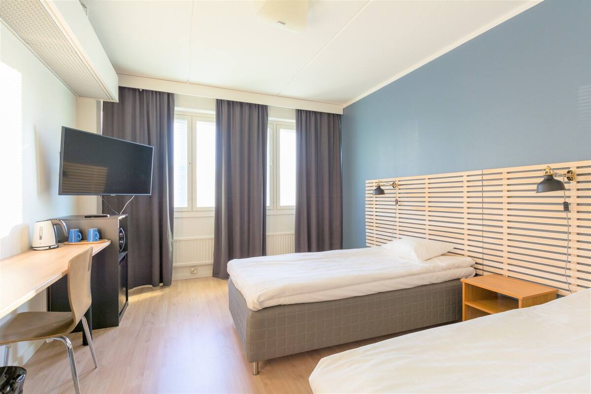 A hotel room with two beds, a desk with a TV, and a window with curtains.
