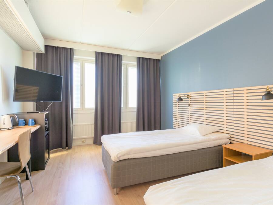 Quarto de hotel com duas camas, uma escrivaninha com televisão e uma janela com cortinas.