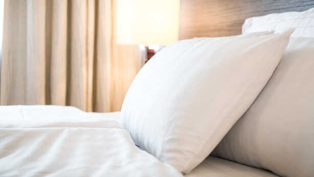Ein ordentlich bezogenes Bett mit weißer Bettwäsche und Kissen, ein Holz-Kopfteil und eine Lampe im Hintergrund.
