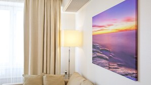 Ein Hotelzimmer mit einem großen Gemälde, das einen Sonnenuntergang über Wasser zeigt, einer Stehlampe und einem beigefarbenen Sofa.