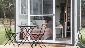 A small, modern cabin with a sliding glass door leading to a bedroom with a bed and a small table. - Saddle Camp Tiny House 2 by Tiny Away (Braidwood)