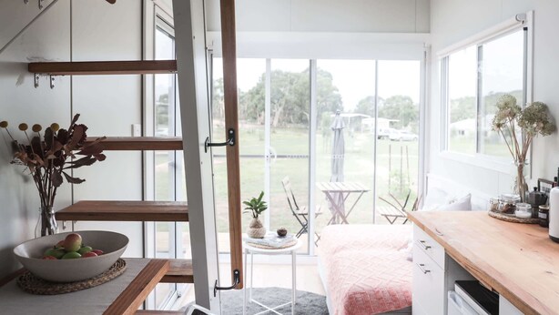 A room with a bed, a wooden table, a bowl of fruit, and a window with a view of the outdoors. - Saddle Camp Tiny House 2 by Tiny Away (Braidwood)