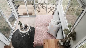 A cozy room with a bed, a small round table with a plant, and a view of outdoor furniture through large windows. - Saddle Camp Tiny House 2 by Tiny Away (Braidwood)