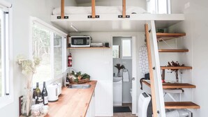 A compact kitchen with a wooden countertop, a microwave, and a sink. There is a small dining area with a chair and a table. - Saddle Camp Tiny House 2 by Tiny Away (Braidwood)