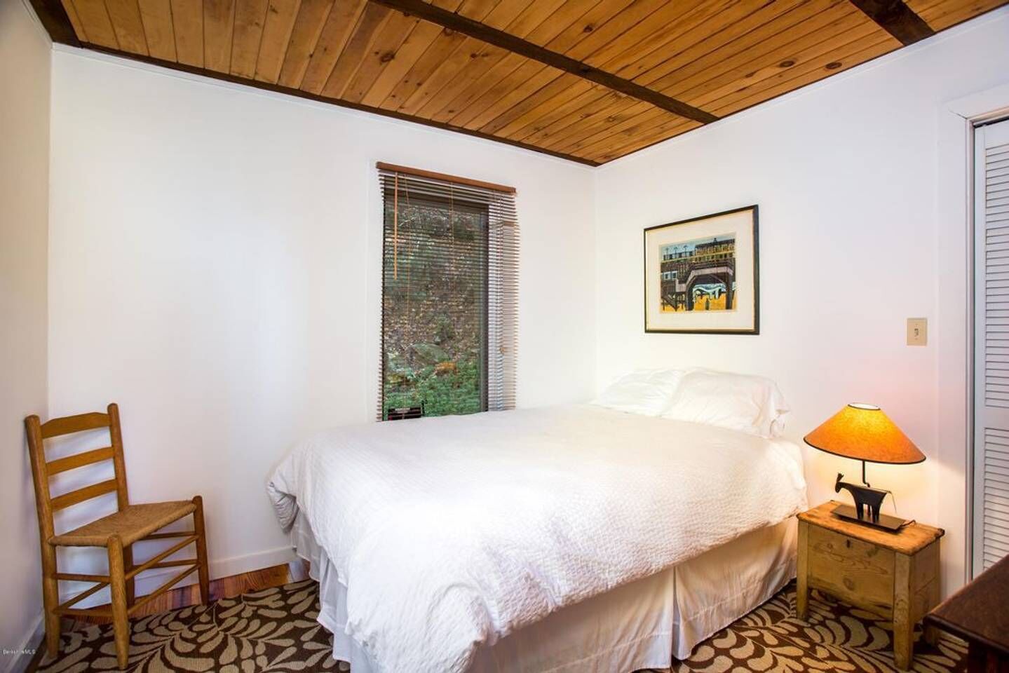 Une chambre à coucher comprenant un lit, une chaise, une table de chevet avec une lampe, une fenêtre avec des stores et un tableau encadré accroché au mur.