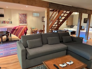 A modern living room with a sectional sofa, a coffee table with remotes, a bookshelf, a painting, and a staircase.