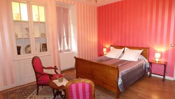 Une chambre à coucher avec un lit en bois, une chaise, une petite table sur laquelle est posé un vase, un lustre et un papier peint à rayures.
