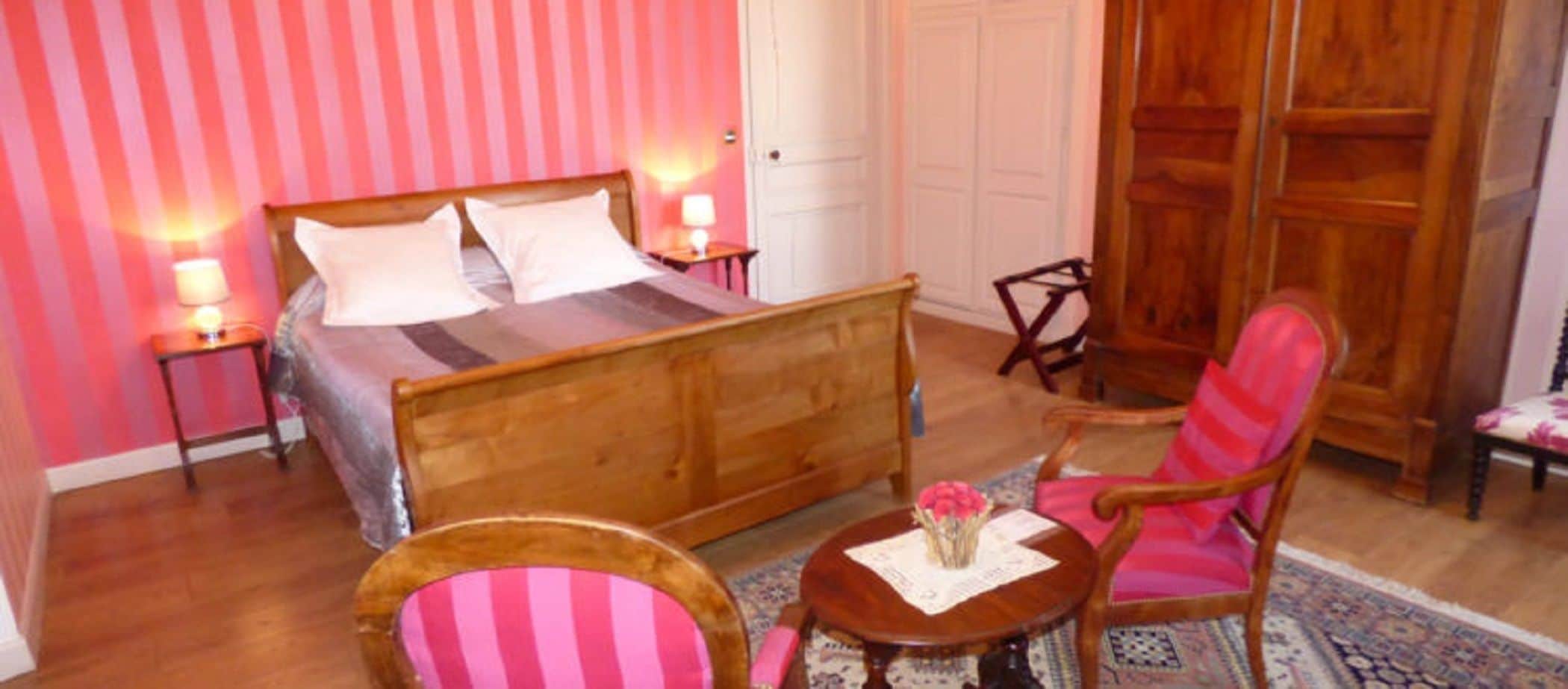 Une chambre avec un lit en bois, deux tables de chevet avec des lampes, une armoire en bois et un fauteuil à rayures roses.