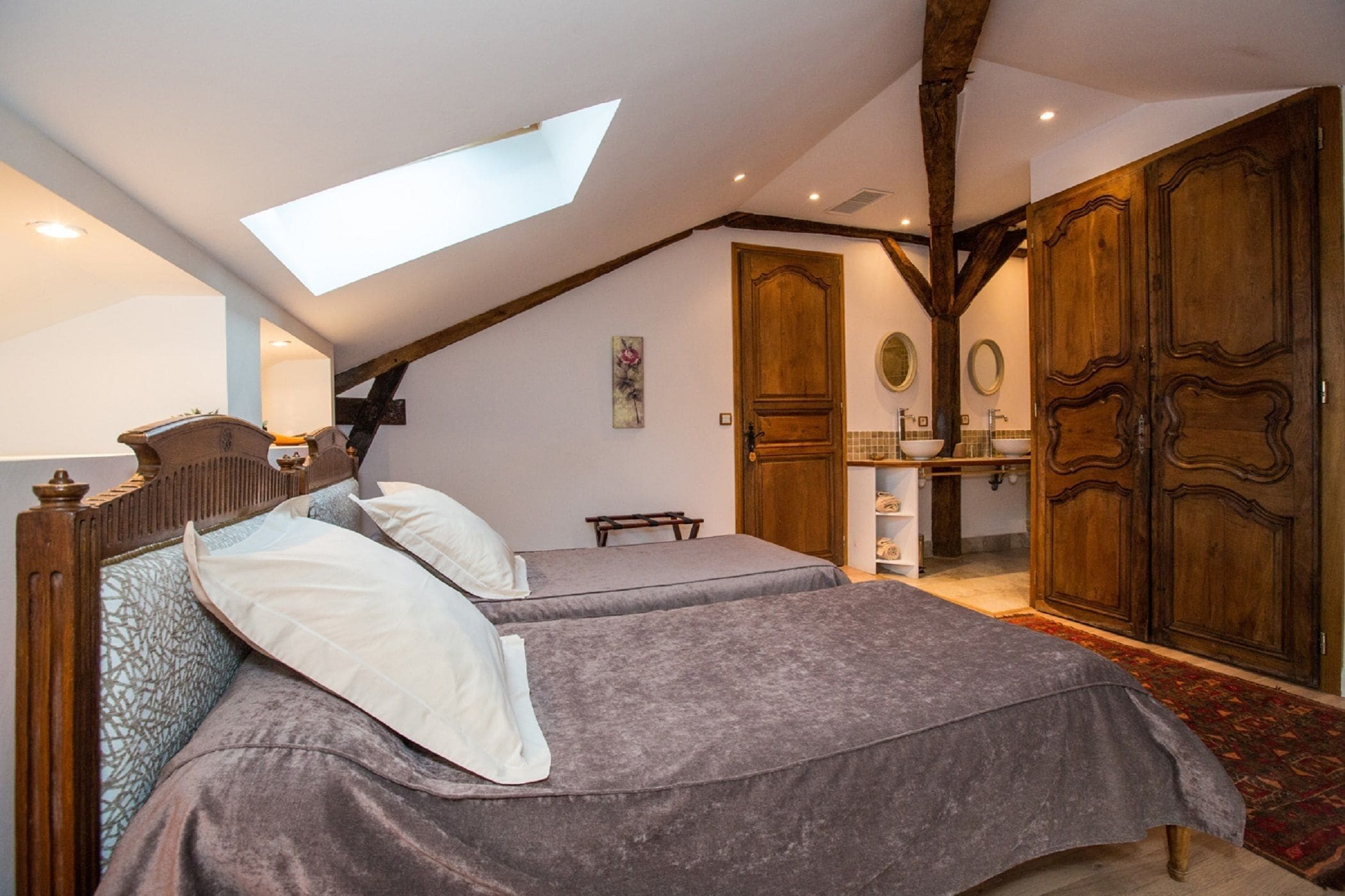 A bedroom with two beds, a skylight, wooden beams, a wardrobe, and a bathroom with a mirror and sink.