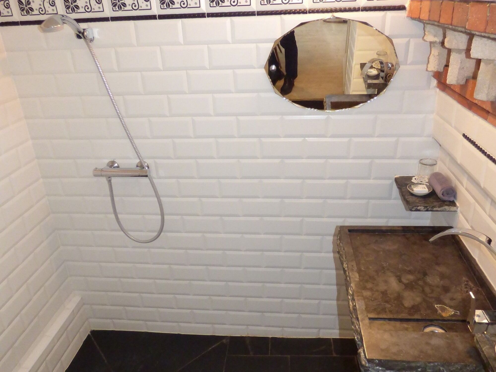 Un baño con paredes de azulejos blancos, ducha con manguera, espejo redondo y lavamanos de mármol con jabonera.