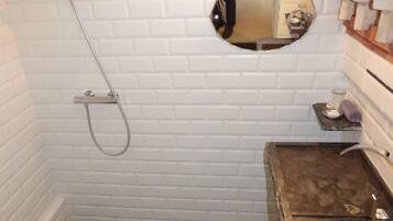 Un baño con paredes de azulejos blancos, ducha con manguera, espejo redondo y lavamanos de mármol con jabonera.