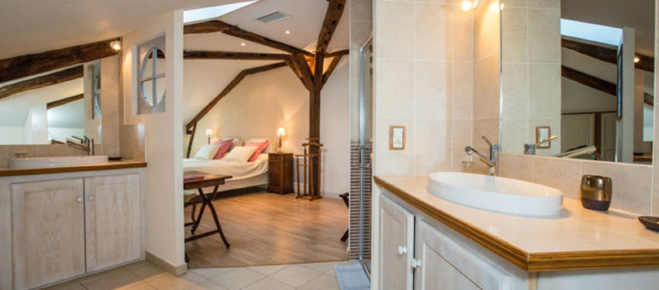 A bedroom with a bed, bedside table, and a window. The room has wooden beams on the ceiling and a large mirror above the bed.