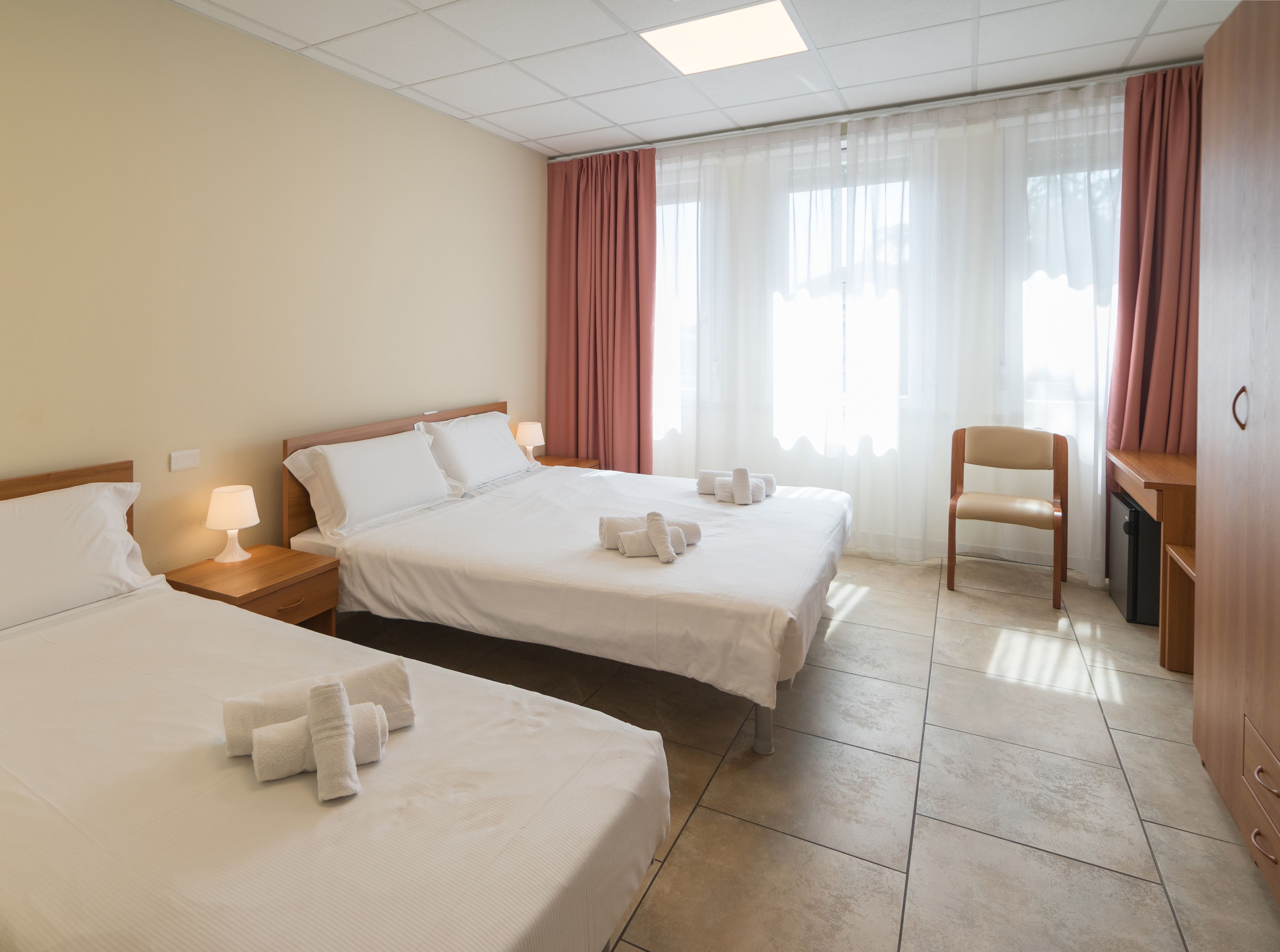 A hotel room with two beds, a desk, a chair, and a window with curtains.
