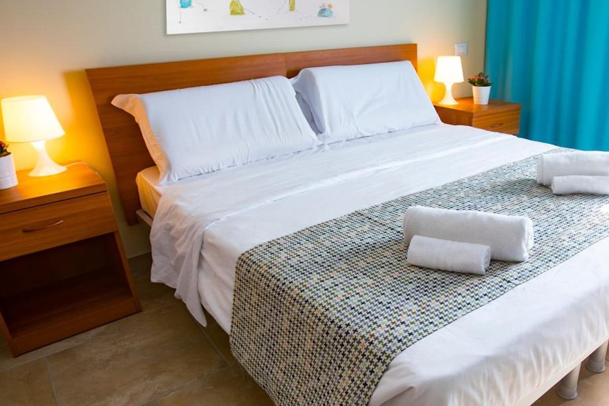 A neatly made bed with white linens, a patterned throw, and two rolled towels on the bed.