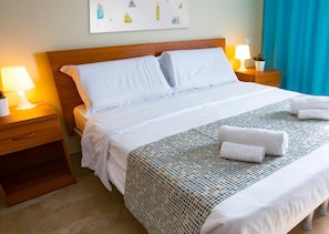 Una cama bien hecha con sábanas blancas, un plaid estampado y dos toallas enrolladas sobre la cama.