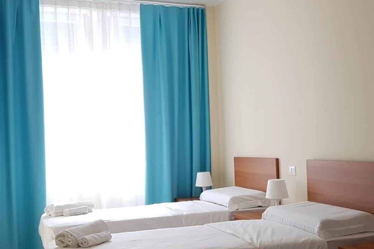 Une chambre d’hôtel avec deux lits simples, une tête de lit en bois, une table de chevet avec une lampe et une fenêtre avec des rideaux bleus.