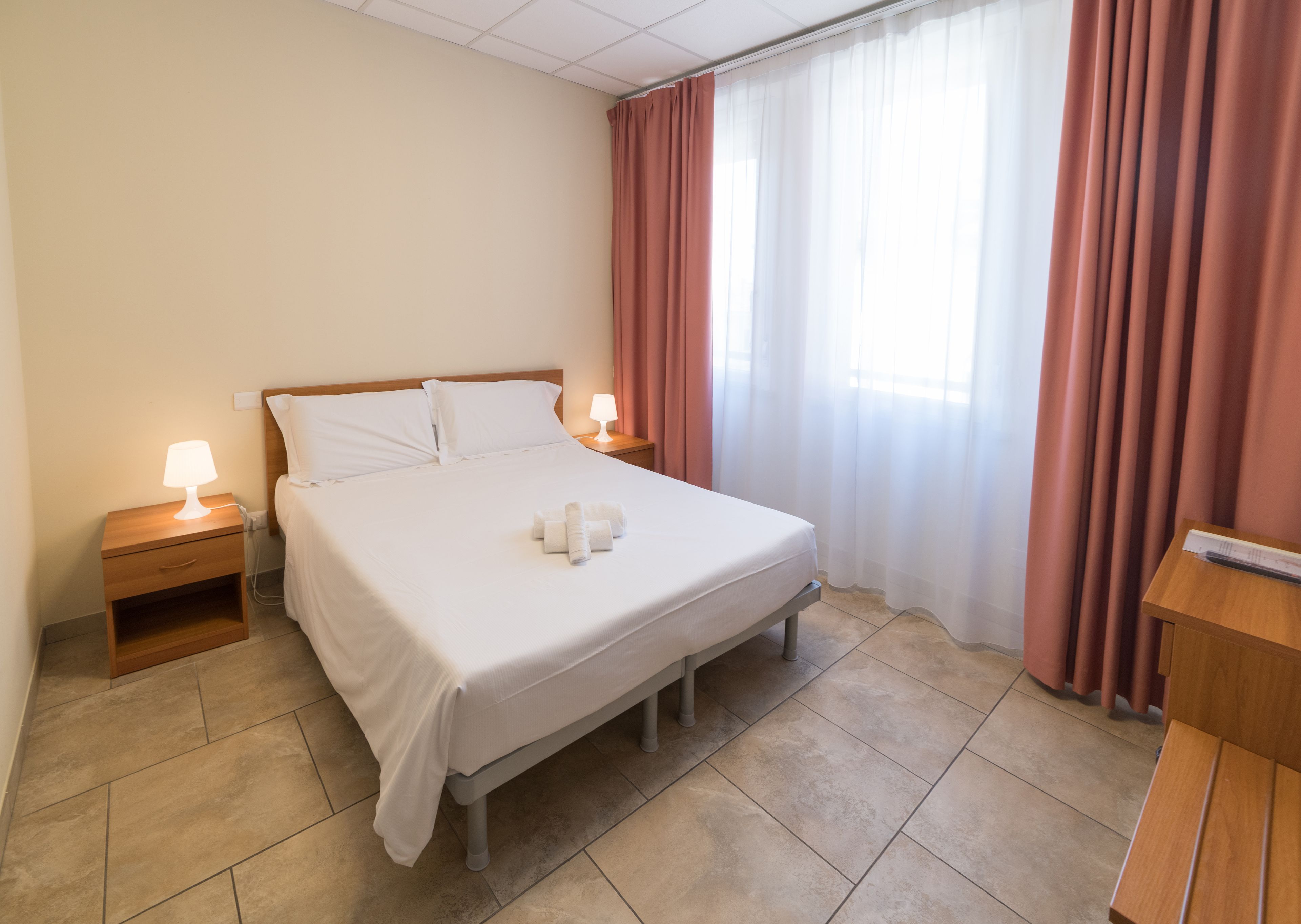 Ein Hotelzimmer mit einem Bett, Nachttischlampen, einem Nachttisch, einer Holzkommode und einem Fenster mit Vorhängen.