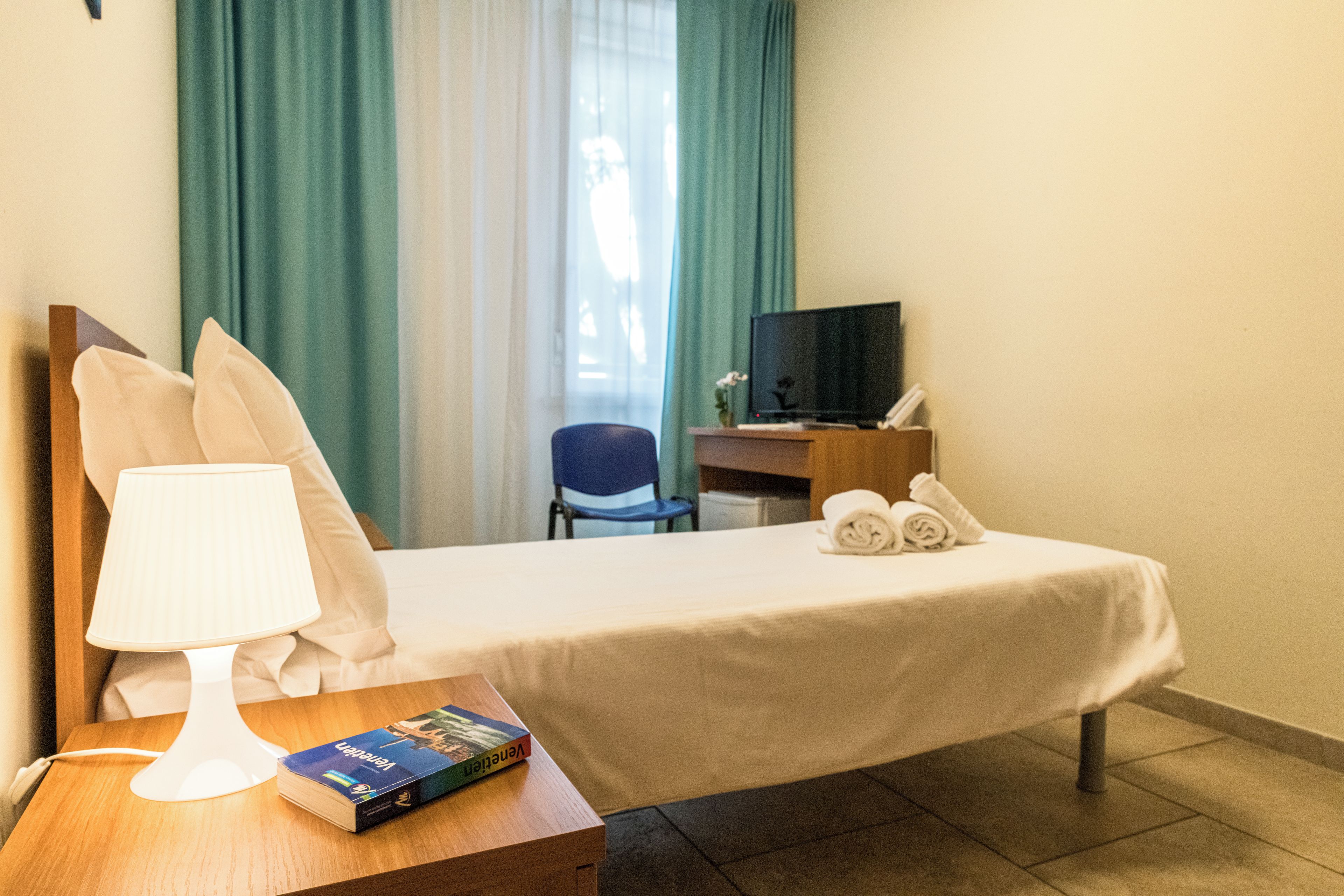 Quarto de hotel com cama, escrivaninha, televisão, abajur e um livro.