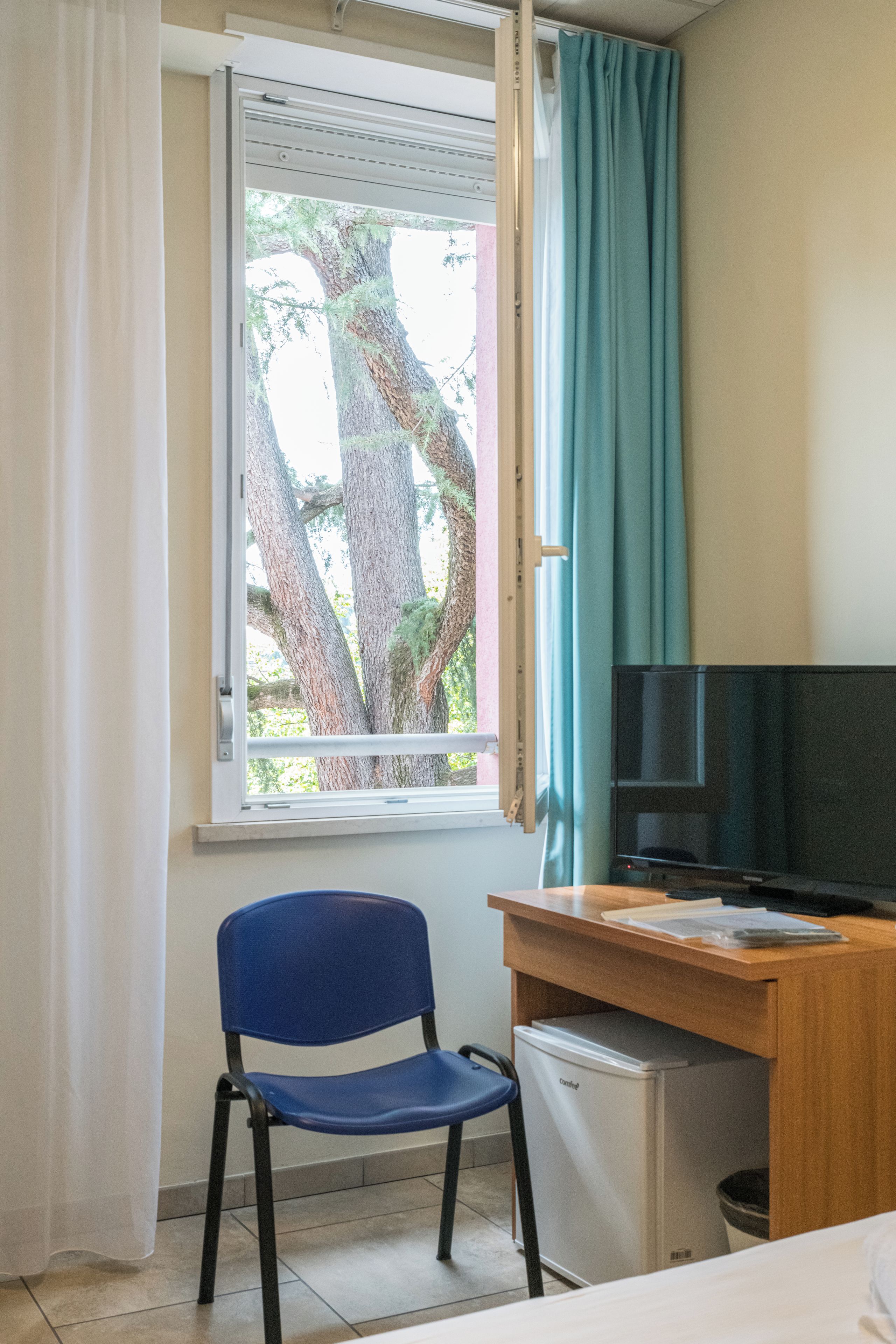 Una stanza con un letto singolo, una scrivania con un computer, una sedia, una televisione e una finestra con tende.